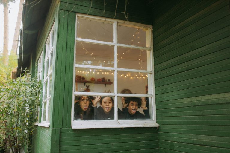 Three joyful children making faces at the window of a cozy cabin, surrounded by autumn scenery.